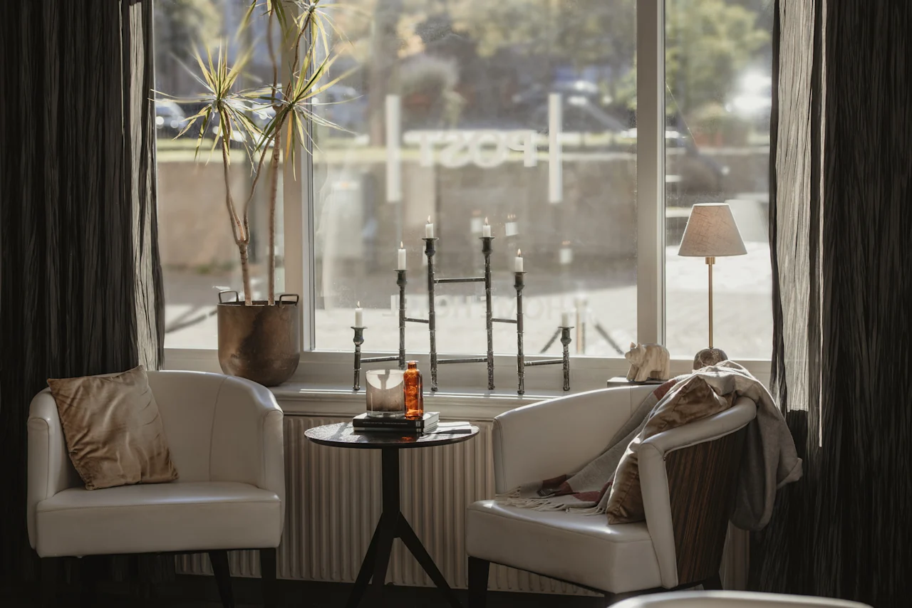 Mysig fönstersittplats med fåtöljer, bord, kandelaber och växt. Text genom fönstret: POST.