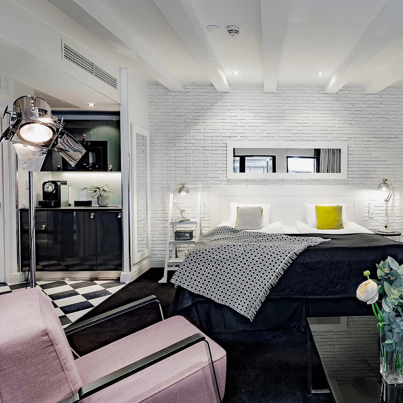 Et moderne soverom har en dobbeltseng med geometriske puter, omgitt av stilige møbler og myk belysning. Et kjøkkenområde og en stilig sofa med blomster gir en koselig atmosfære.