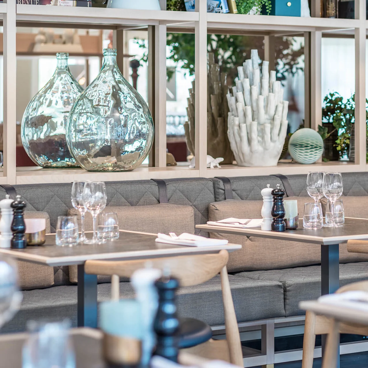 Restaurangbord är dukade med glas och servetter på gråa vadderade bänkar. Bakom pryder dekorativa glasflaskor och växter hyllor av trä och skapar en modern och luftig atmosfär.