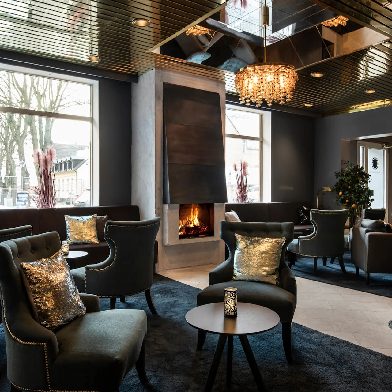 Ett loungeområde har eleganta stolar runt bord, en öppen spis med modern kåpa och stora fönster. Mjuk belysning och dekorativa kuddar skapar en mysig atmosfär.