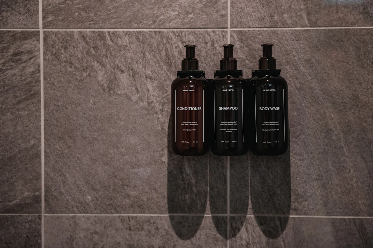 Tre mørke flasker merket balsam, sjampo og kroppsvask er montert på en teksturert grå flislagt vegg på et bad.