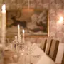 Stearinlys oplyser et formelt dækket spisebord med glas og servietter, omgivet af polstrede stole. Et maleri af en ren pryder den mønstrede væg, med bløde lysarmaturer.