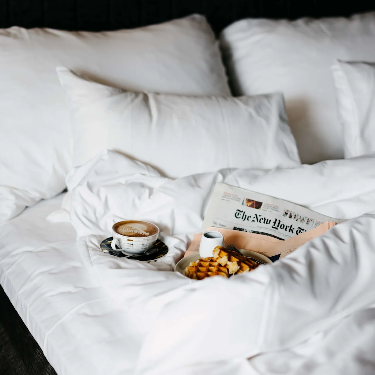 Et frokostbrett med kaffe og vafler står på en seng, sammen med avisen "The New York Times", omgitt av fluffy hvite puter og sengetøy.