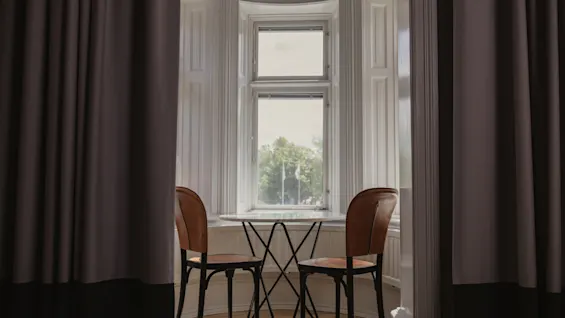 Ett litet bord med två stolar står vid ett burspråk, inramat av gardiner, med utsikt över träd utomhus.
