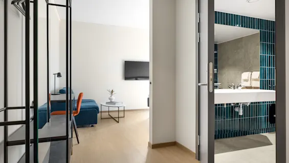 Et moderne hotellrom med en blå sofa og en oransje stol, med utsikt inn i et bad med blå fliser.