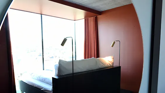 Seng med hvide puder, indrammet af to lamper, reflekteres i et rundt spejl. Gulv-til-loft-vinduer og brændte orange vægge skaber en moderne, minimalistisk rumstemning.
