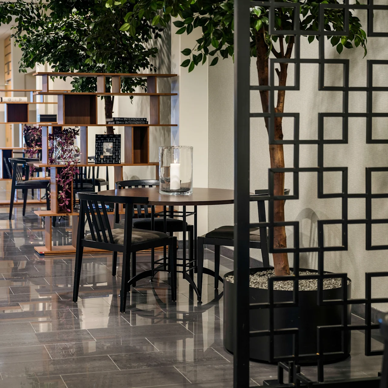 Tables and chairs line a corridor with potted trees and decorative screens. The setting appears to be a modern indoor space with shiny tiled floors and a bright, airy atmosphere.
