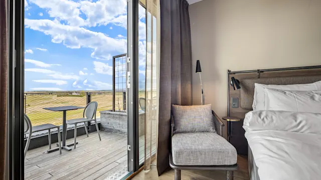 Et koselig hotellrom med en seng og lenestol; glassdører åpner seg mot en balkong med stoler og bord, med utsikt over et stort gresslandskap under en delvis skyet himmel.
