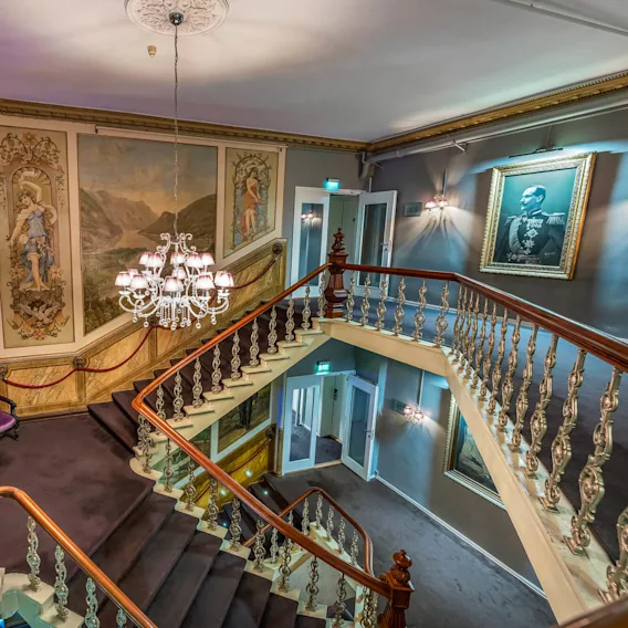 Grand staircase curves upward, adorned with ornate paintings and a central chandelier, set in a luxurious, art-filled interior featuring a purple bench and framed portrait on the wall.