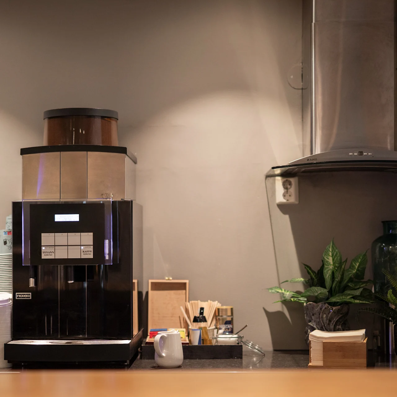 En kaffemaskin står på en disk, omgiven av engångskoppar och krukväxter. I närheten är en köksfläkt fäst vid väggen. Miljön verkar vara ett café.