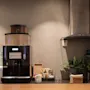 En kaffemaskin står på en benk, omgitt av engangskrus og potteplanter. I nærheten er en avtrekkshette festet til veggen. Omgivelsene synes å være en kafé.
