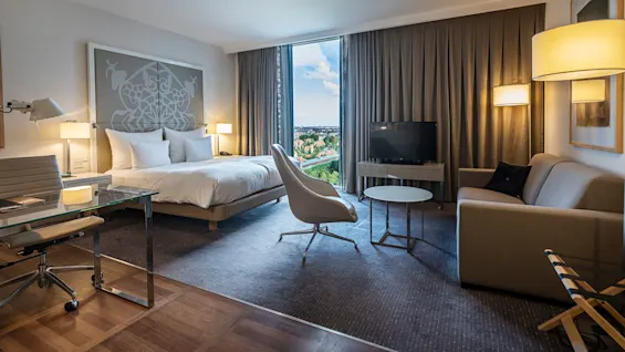Ett modernt hotellrum med en stor säng prydd med ett mönstrat sänggavel. Ett skrivbord av glas, stol och soffa finns i närheten med utsikt över hustak och träd.