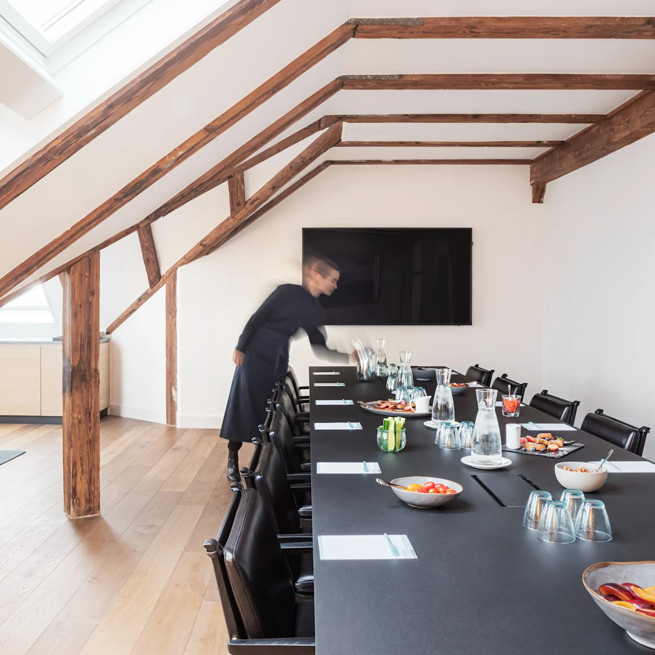 A conference table is set with notepads, glasses, and snacks, while a person arranges items. The room has wooden beams, a wall-mounted screen, and slanted windows.