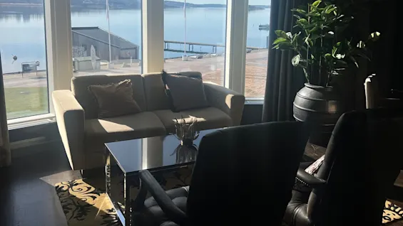 A sofa and chairs are arranged around a glass table in a sunlit living room, with large windows overlooking a calm, scenic waterfront.