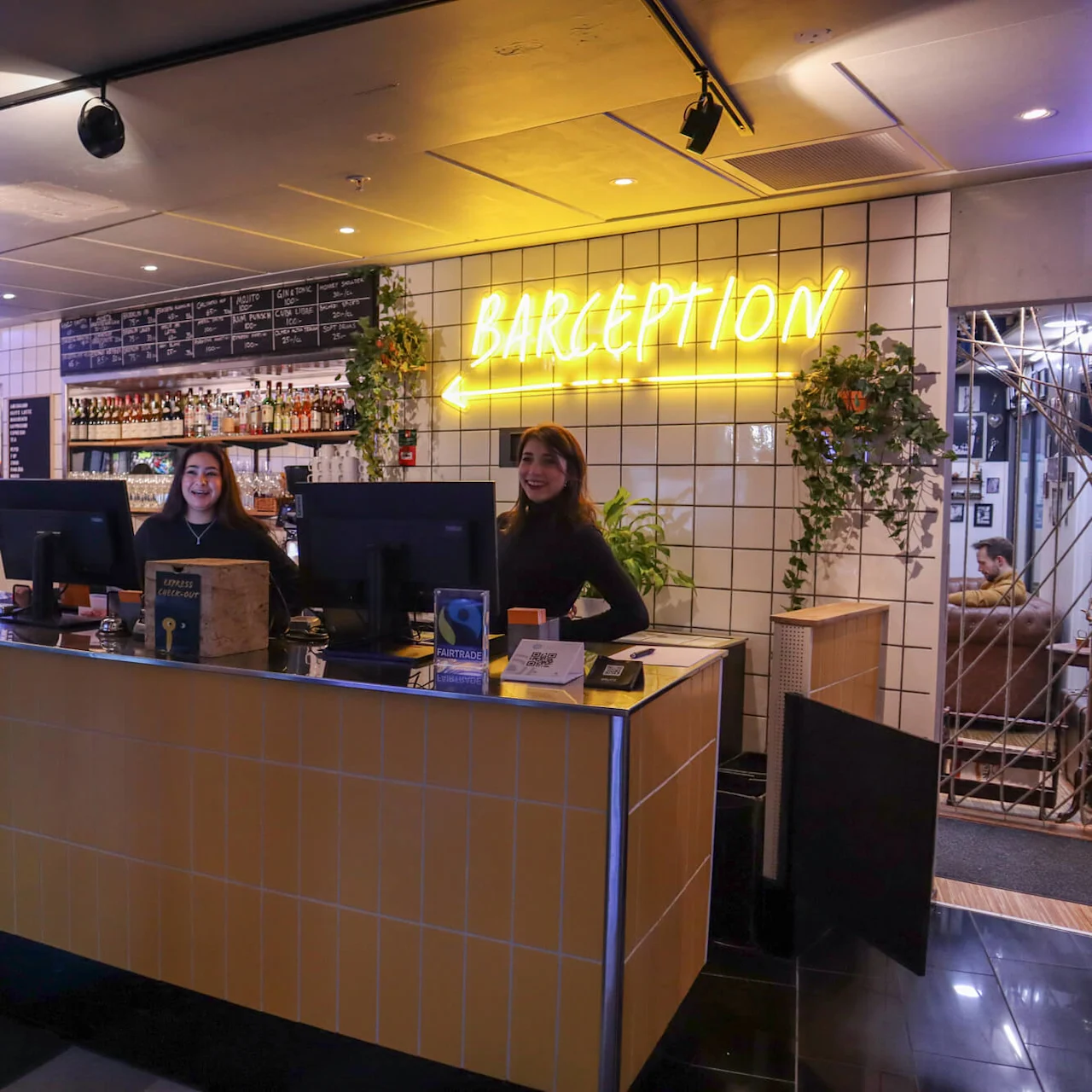 Two staff members stand behind a hotel reception desk labeled "BARCEPTION," smiling in a warmly lit interior with bar elements.