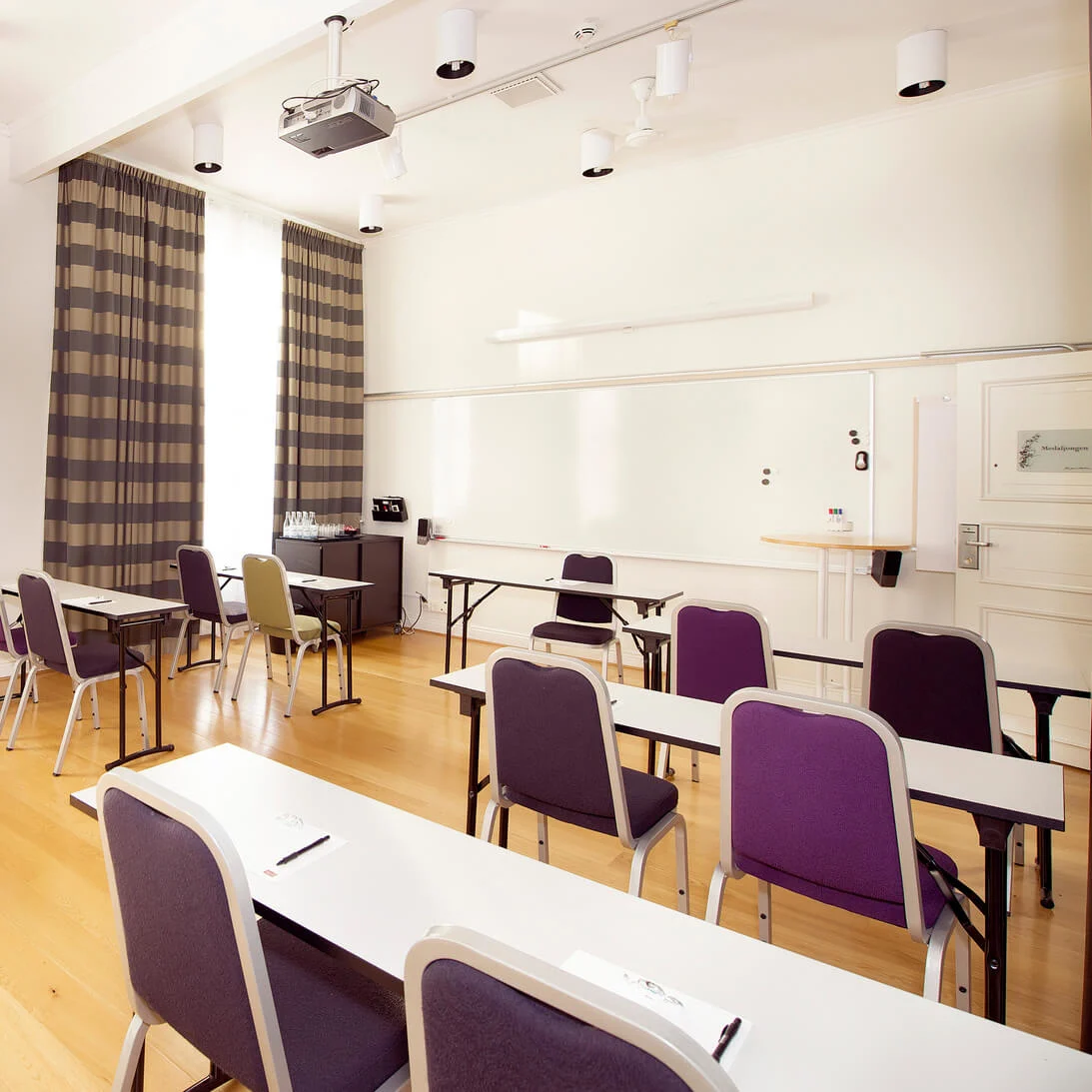 Chairs and tables arranged in an empty classroom with curtains, a whiteboard, and a projector.