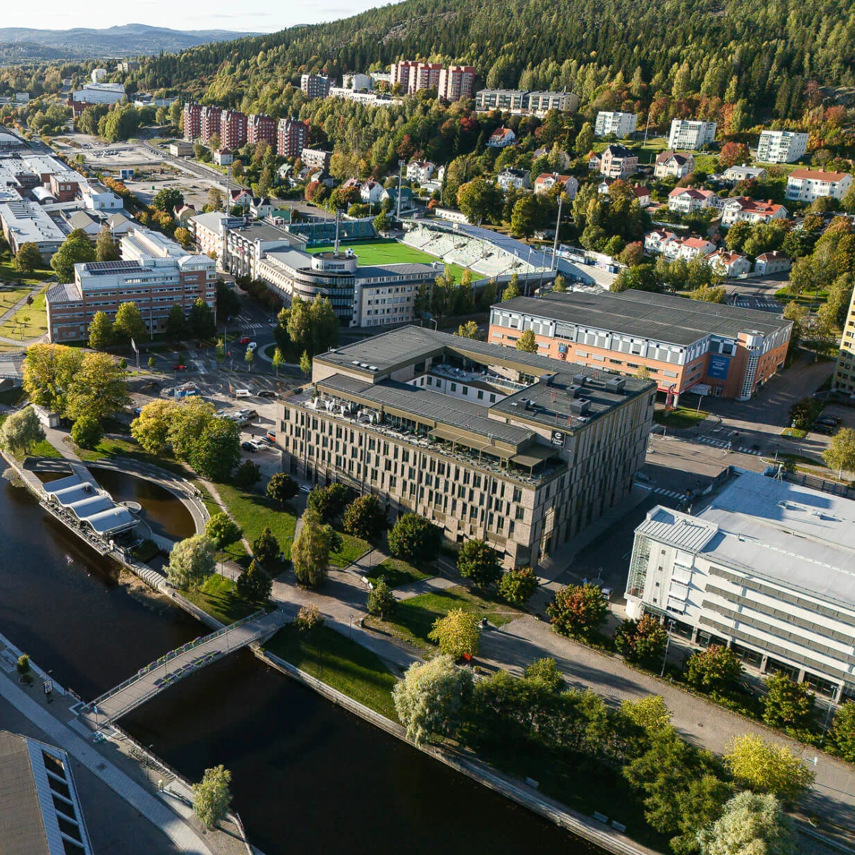 Luftfoto af bybillede med en flod, der løber gennem centrum med flere broer. Omkringliggende område inkluderer bygninger, veje og en grøn skovklædt bakke.