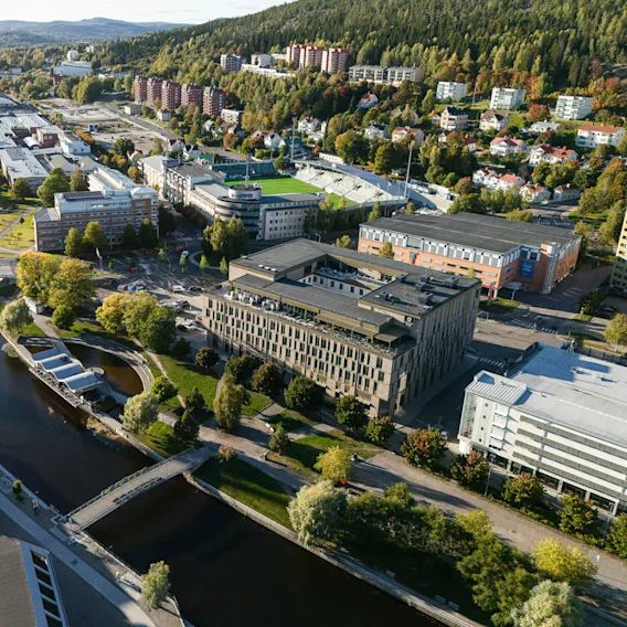 Aerial view of a cityscape, with a river running through the center featuring multiple bridges. The surrounding area includes buildings, roads, and a green forested hillside.