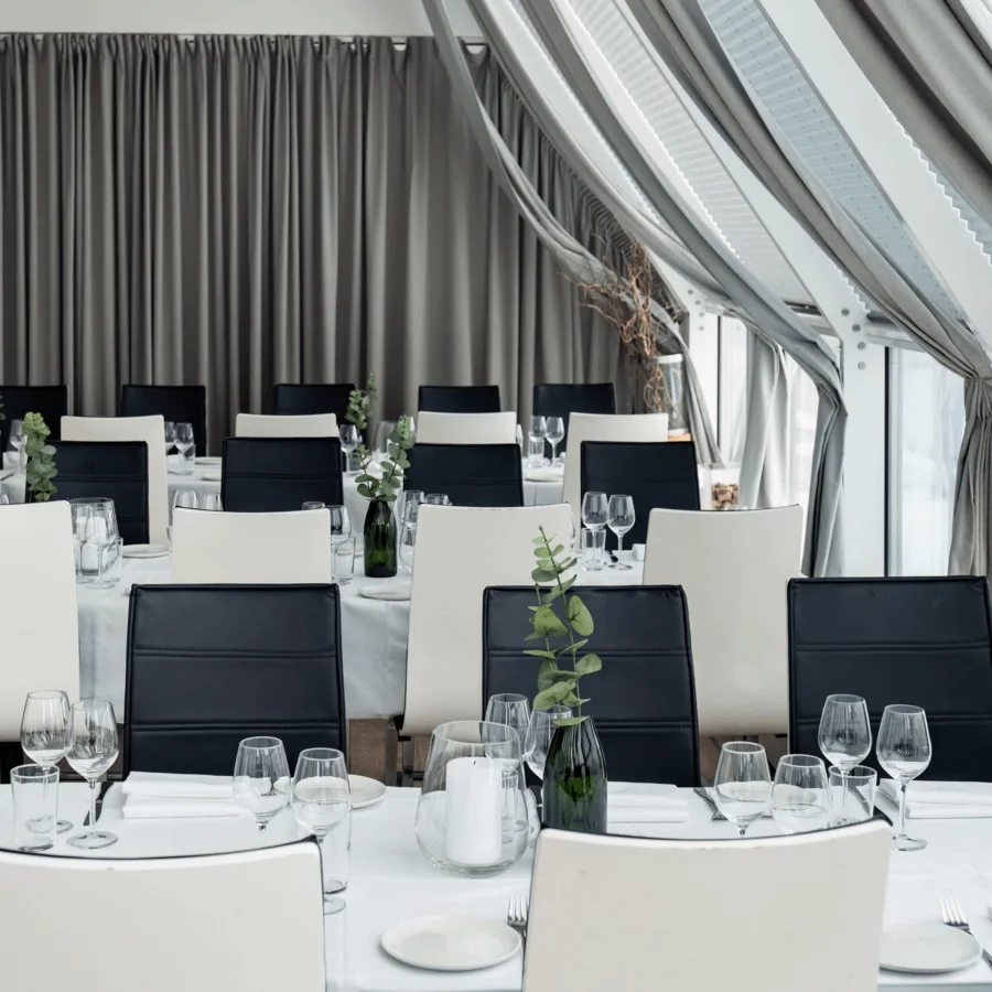 Bord dukade med vita dukar och glas i en restaurang, med svarta och vita stolar, gröna växter i vaser, omgivna av beige gardiner och stora fönster.