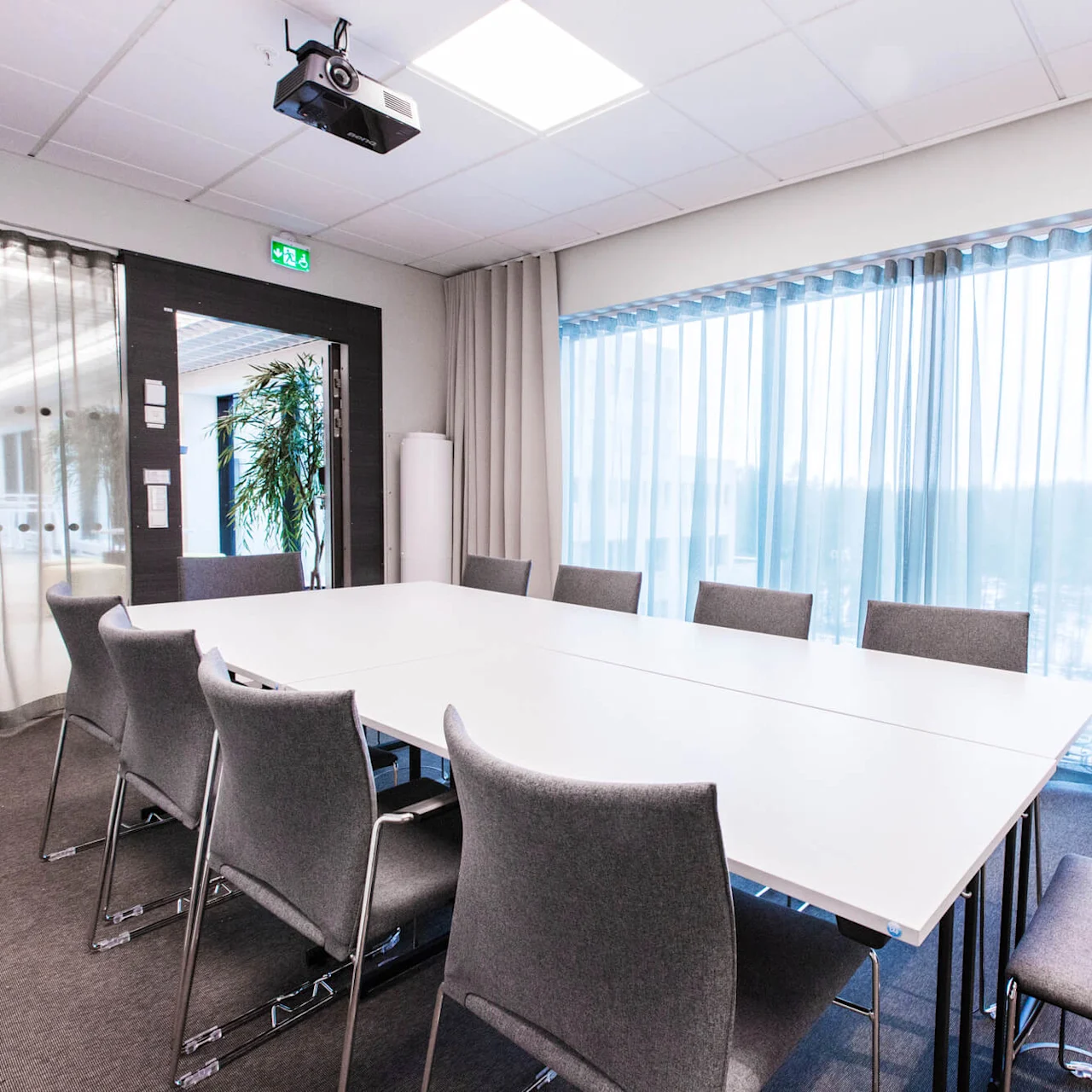 Ett stort, vitt bord omgivet av grå stolar i ett väl upplyst konferensrum. En projektor hänger i taket, och skira gardiner täcker stora fönster på ena sidan.