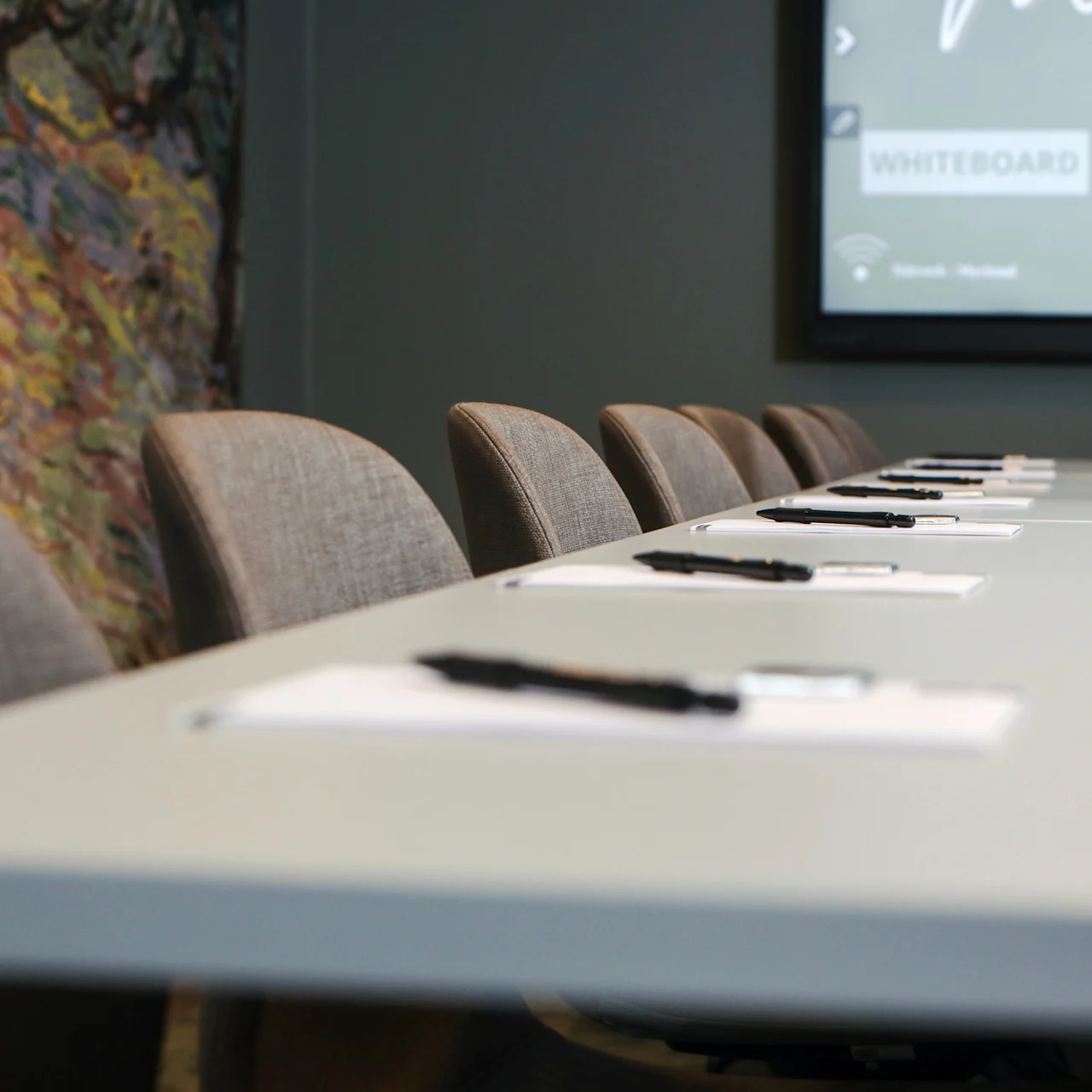 Meeting room table with notepads and pens aligned on top, surrounded by padded chairs. A wall screen displays "WHITEBOARD WEBBLÄSARE", next to a colorful abstract mural.