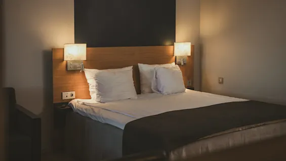 A neatly made double bed with white pillows and sheets is illuminated by warm wall-mounted lamps on a wooden headboard in a cozy, dimly lit bedroom.