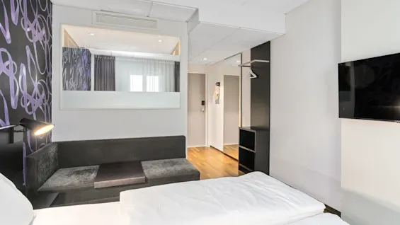 A modern hotel room with a bed, couch, and mounted TV, featuring a decorative wall and a wooden floor.