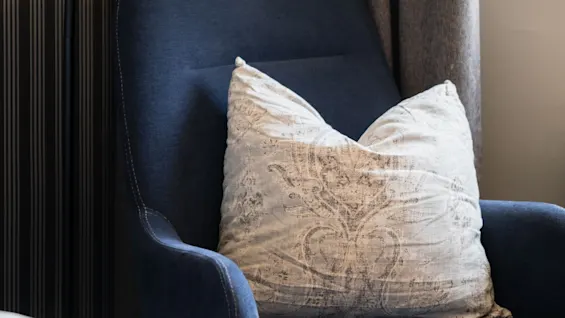 Et hyggeligt hjørne i et værelse med en mørkeblå lænestol, en mønstret pude, en gulvlampe og en seng.