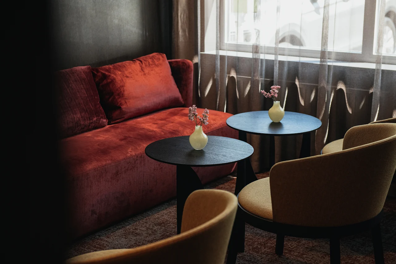 Innbydende hotell sittegruppe med en plysj rød sofa, to små bord med blomsterdetaljer, og komfortable stoler ved et lyst vindu.