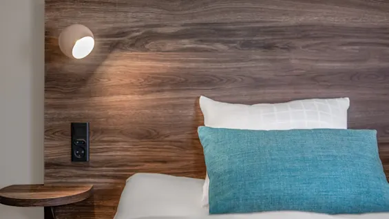 A bed with white and blue cushions rests against a wooden headboard featuring a sconce light and small wooden shelf, creating a cozy and minimalistic bedroom setting.