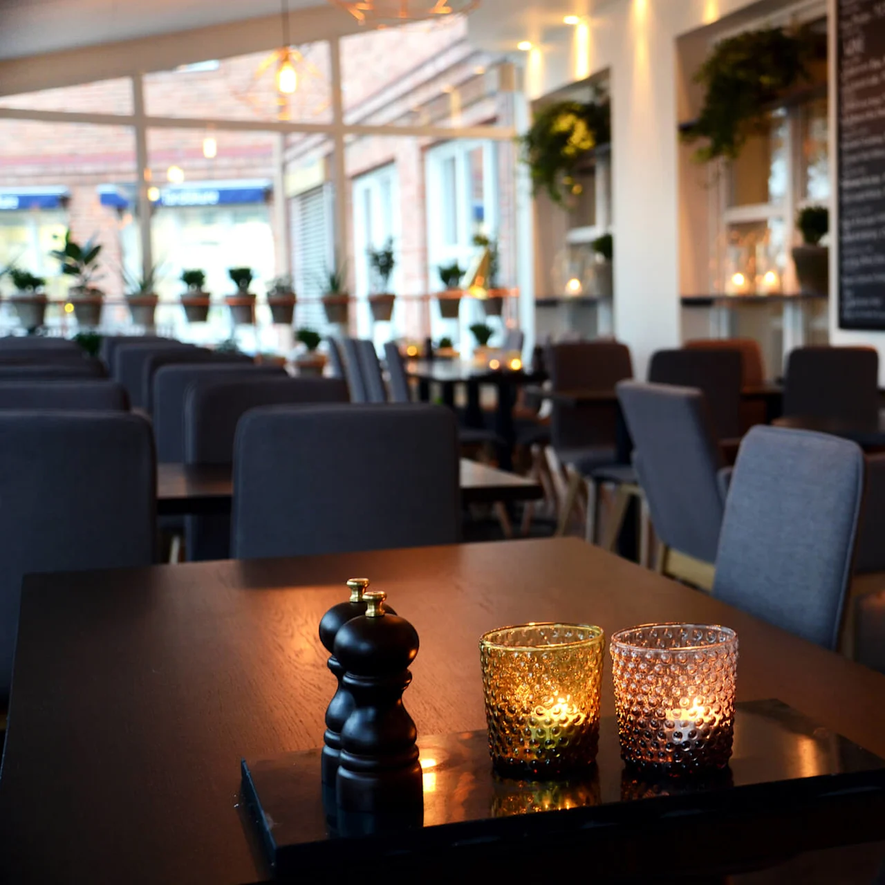 A restaurant interior with dim lighting, featuring tables set with candles, surrounded by empty gray chairs, and a wall of wine shelves in the background.