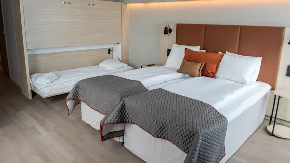 Moderni hotellihuone, jossa kaksi sänkyä ja alaslaskettava lisävuode, tarjoten joustavat nukkumisjärjestelyt.