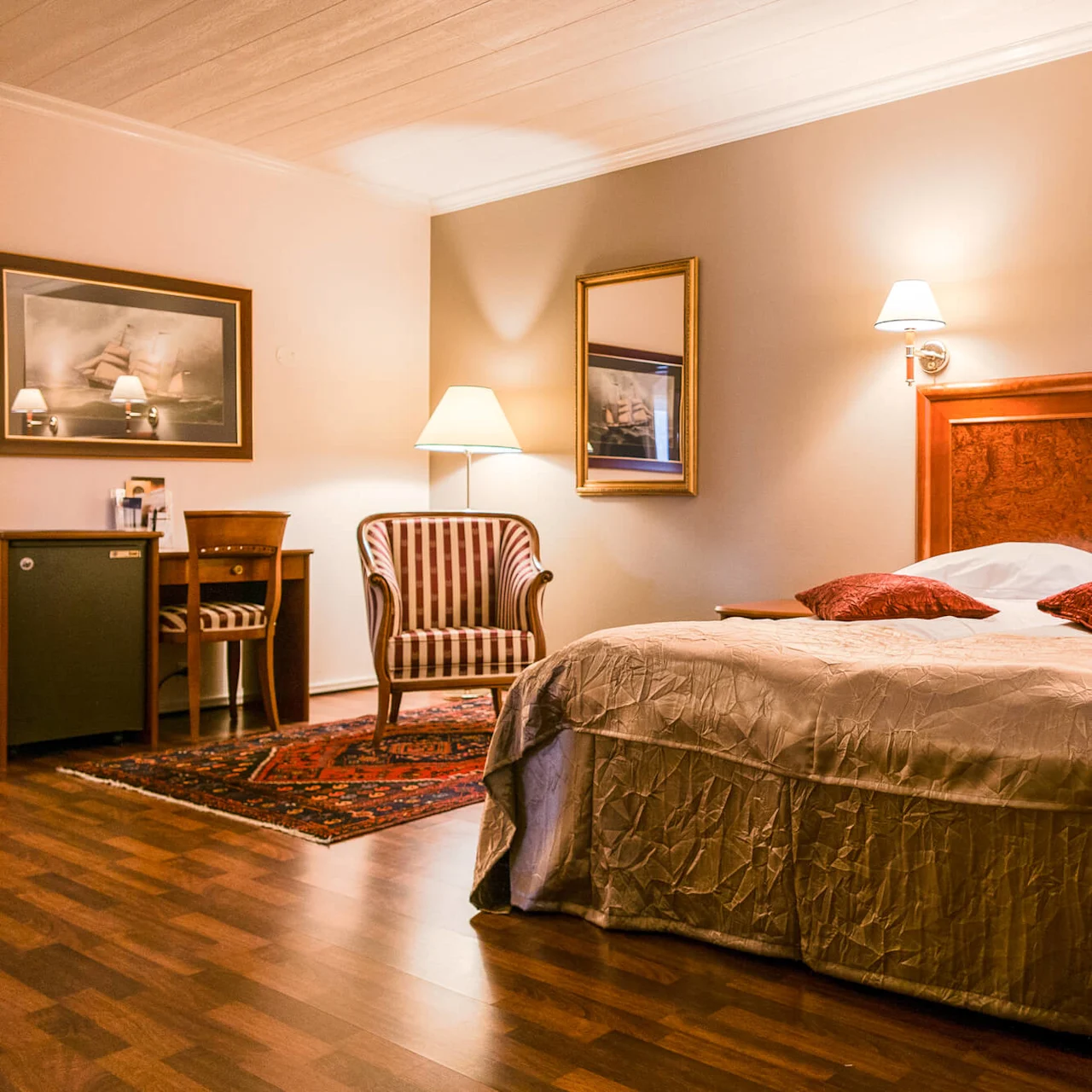 A bed with two red pillows sits in a warmly lit hotel room. Nearby, a striped chair and a small desk are positioned on a patterned rug.