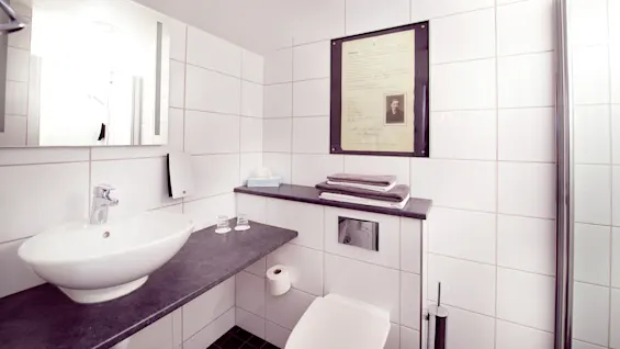 A modern bathroom with white tiles, featuring a sink, mirror, framed document; towels on a shelf add a touch of comfort.