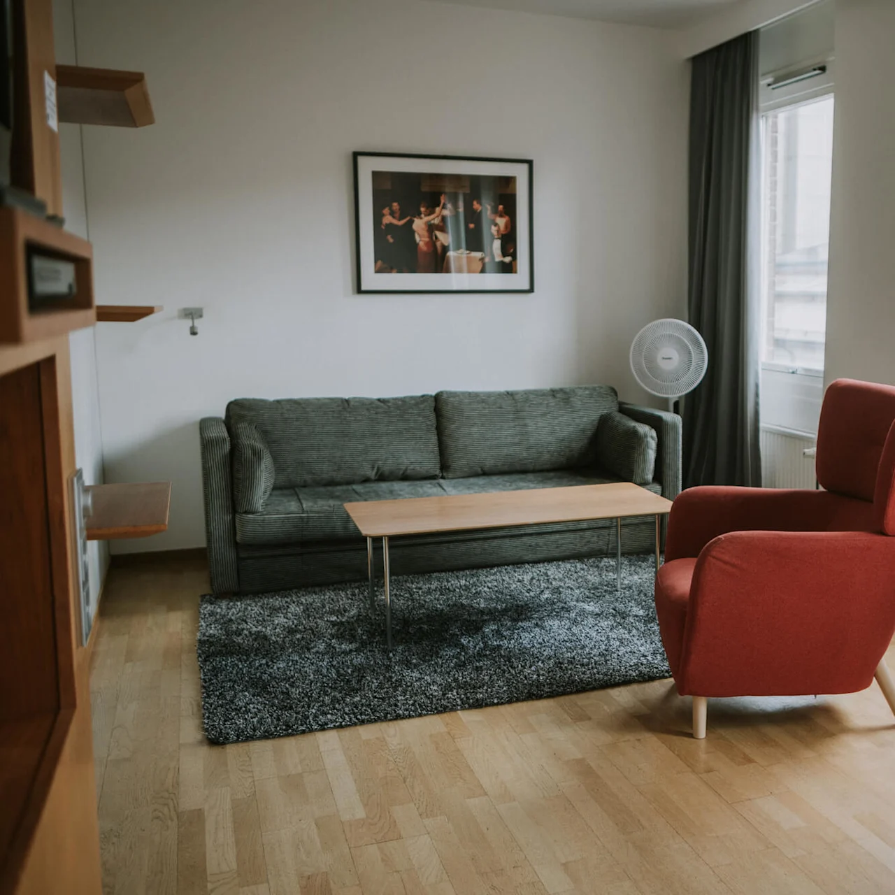 En rød lænestol vender mod en grøn sofa i et lyst rum med trægulv, sofabord og et indrammet fotografi på væggen. Store vinduer lader naturligt lys komme ind.
