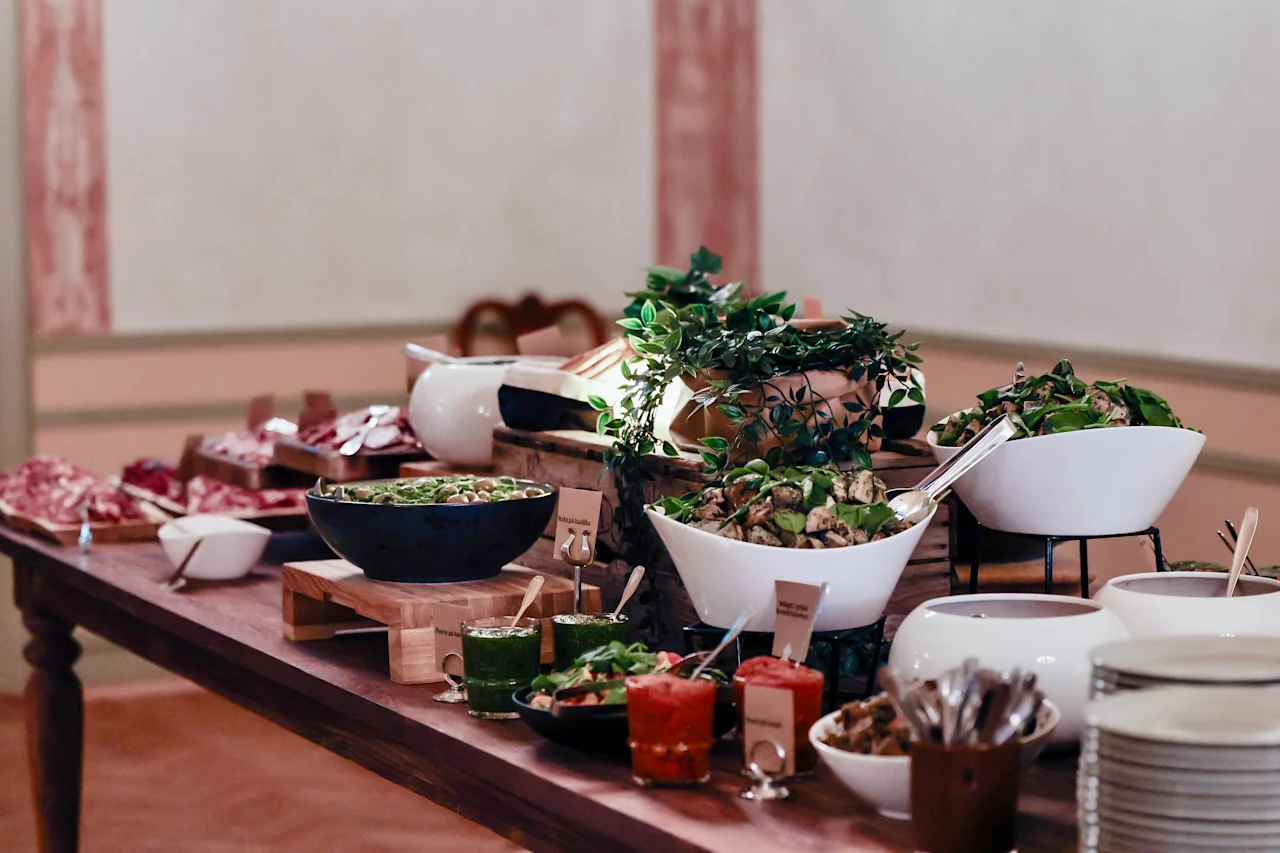 A buffet table features assorted dishes in white bowls, including salads and sauces, accompanied by serving utensils. It is set in an elegant dining room with pink-accented walls.