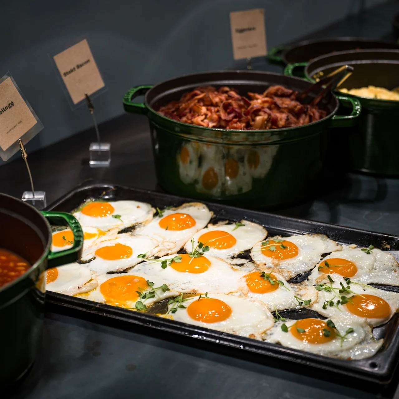 Spejlæg på en bakke i en buffet, omgivet af gryder med bacon og bagte bønner. Skiltene lyder: "Speilegg" og "Stekt Bacon".