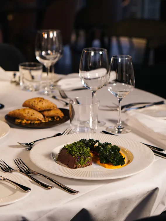 En tallerken med gourmetmat pyntet med grønt og saus står på et dekket bord med vinglass og brød ved siden av i et restaurantmiljø.