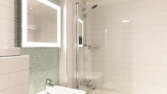A modern bathroom at Home Hotel Grand Bodø featuring a sink, an illuminated mirror, and a shower over a bathtub.