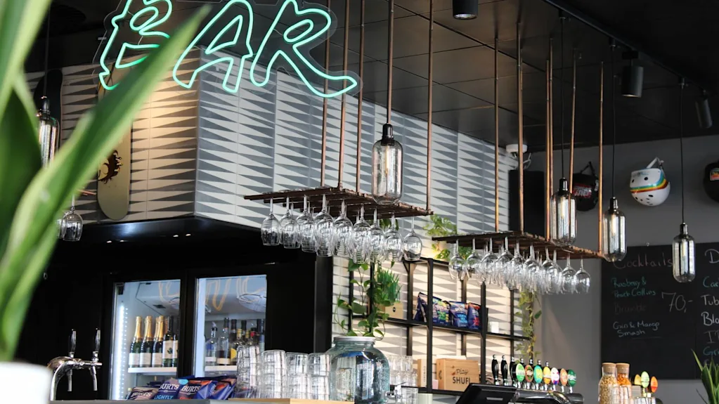 A modern bar with a hanging system for wine glasses and decorative lamps in an industrial style. Above the bar, a green neon sign displays the word "BAR."