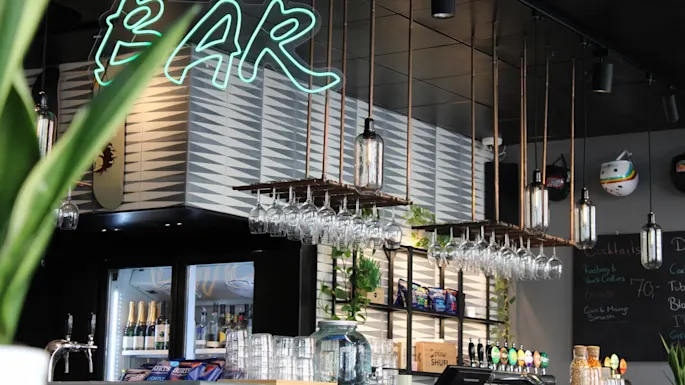 A modern bar with a hanging system for wine glasses and decorative lamps in an industrial style. Above the bar, a green neon sign displays the word "BAR."