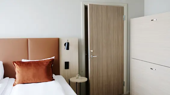 Moderni hotellihuone siististi pedatulla sängyllä, yöpöydällä ja osittain avoimella ovella, mikä viittaa kutsuvaan tilaan.