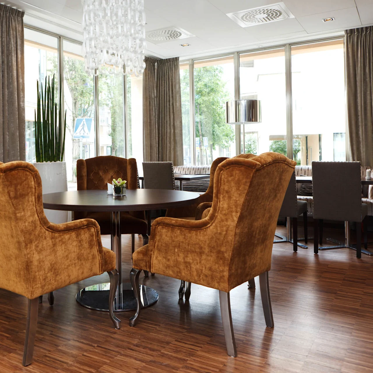 Velvet chairs surround a round dining table in a well-lit, modern room with large windows and decorative indoor plants.