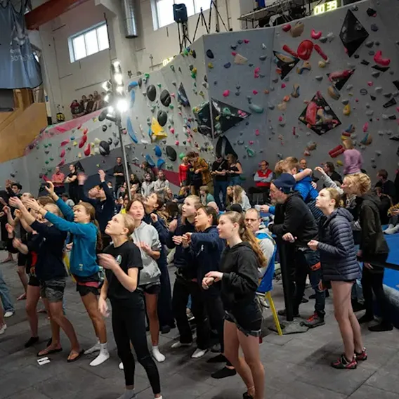 En folkmassa samlad i en inomhusklätterhall, tittar upp mot en klättervägg och följer en aktivitet.