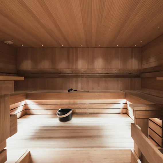 A wooden sauna interior with tiered benches and dim lighting; a black bucket rests on the floor, and a ladle is placed on a bench.