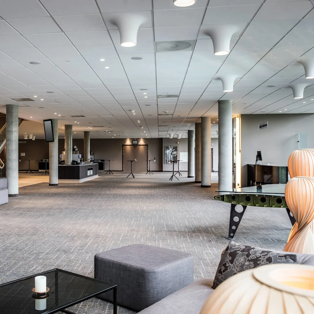 Romslig lobby med grå sofaer og bord, to spirallamper og potteplanter. Teppelagte gulv fører til en trapp og resepsjonsområde med taklys.
