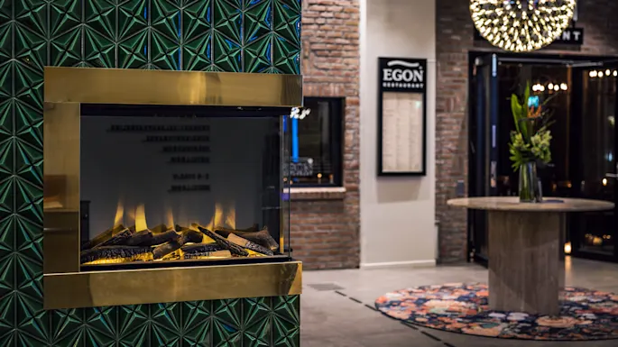 Sähköinen takka realistisilla liekeillä vihreällä kuvioidulla seinällä. Taustalla tiiliseinä ja kyltti jossa lukee "EGON RESTAURANT". Pyöreä pöytä kukkien kera koristeellisen kattokruunun alla.