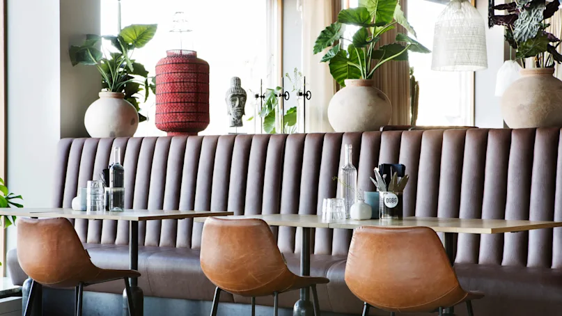 Seating area with brown leather chairs and a wooden table, adorned with glassware and plants. Decorative items like vases and a red lantern are spaced along a cushioned bench.