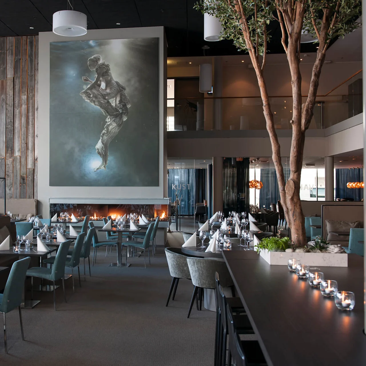 Bord med hvite servietter er dekket i en moderne restaurant med et stort abstrakt maleri. Trær og glassdekor skaper en stilfull atmosfære. Synlig tekst: "KUST".