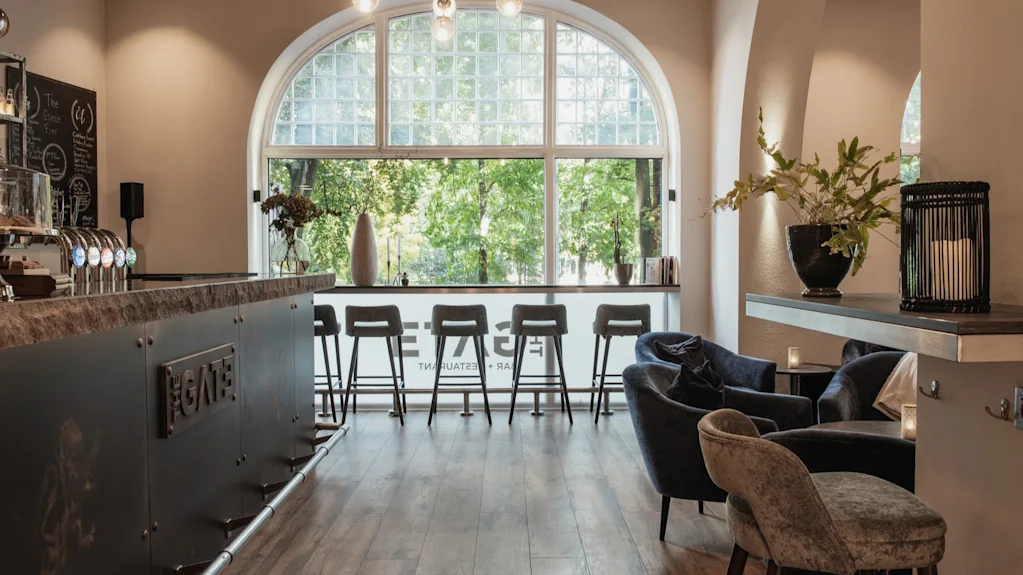 Interiør af en bar og lounge med et stort buet vindu, der viser grønne træer. Barstole og lænestole. FGATE RESTAURANT.